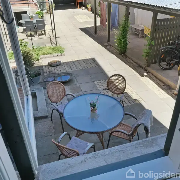 Et bord med fire stole står på en terrasse. Bordet har en lille plante på toppen. Der er en cykel og nogle potteplanter i baggrunden.