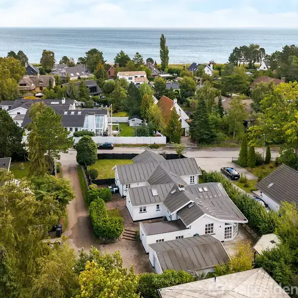 Et parcelhus med gråt tag står midt i en frodig boligområde. Træer omgiver kvarteret, og i baggrunden ses havet, hvilket skaber en rolig og idyllisk atmosfære.