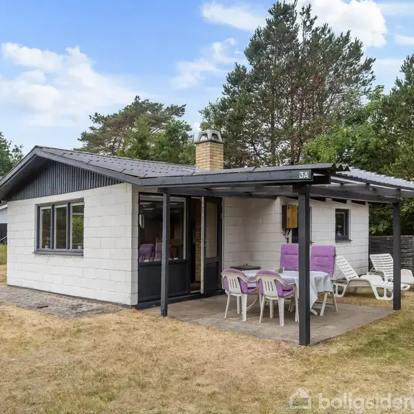 Et lille sommerhus med hvid facade og skråt tag, står stille omgivet af træer og en græsplæne. En terrasse med lilla stole og et bord er under et halvtag. Husnummeret er 53A.