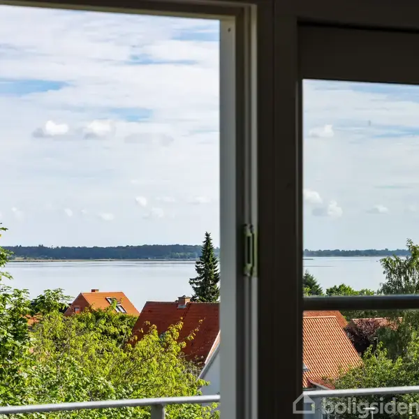 Et vindue med udsigt over rødt tagede huse i en frodig grøn have, baggrund med vand og en skovklædt kystlinje under en let overskyet himmel.