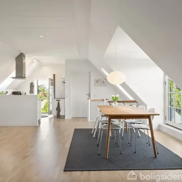 En lys stue med trægulv. Reol fyldt med bøger står til højre, mens et gammelt ur og en kommode med lampe står til venstre. Grå tæppe i midten.