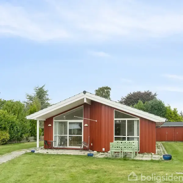 Rødt, enkelt hus med hvidt tag står stille på en grøn græsplæne. Havebord og stole ses foran huset. Omgivet af tæt bevoksning mod en blå himmel.