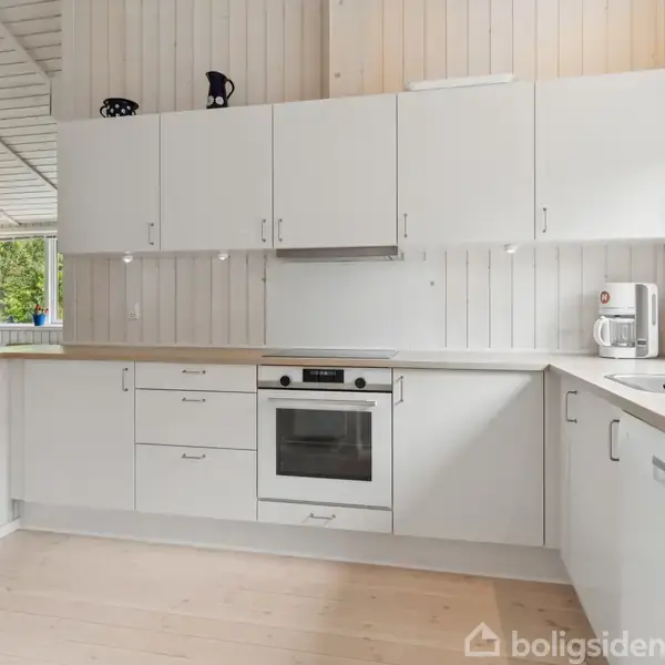 Et hvidt køkken med skabe monteret på væggen og en indbygget ovn i midten. Til venstre ses en åben dør til et andet rum. Rummet er lyst.