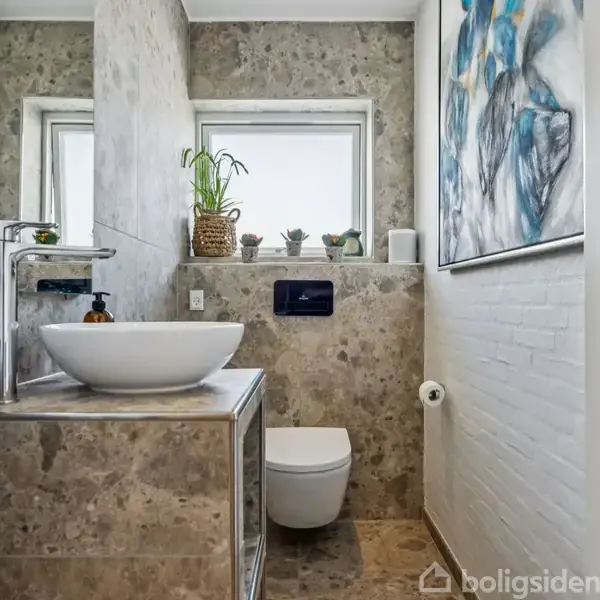 En moderne håndvask på et marmorbord med en hvid porcelænsskål, står ved siden af et væghængt toilet i et stilfuldt badeværelse. To vinduer og en stor kunstmaleri pryder væggen.