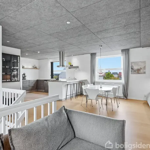 Stue med grå sofa. Køkkenområde til venstre med ø, barstole og sort skab. Spisebord med stole foran vindue. Lyse trægulve og grå lofter. Udsigt til udenfor.
