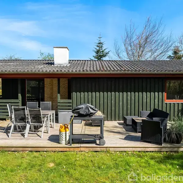 Et sommerhus med en træterrasse er omdrejningspunktet. Terrassen har havemøbler, en grill og planter. Omgivet af grøn have under en blå himmel, skaber det en afslappet atmosfære.