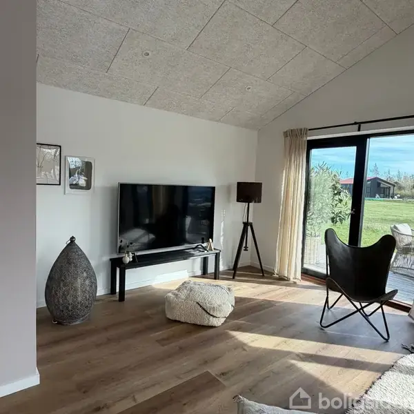 TV på væggen står på et stativ i en lys stue med trægulv. Omkring er der en puf, en gulvlampe, en lænestol og store vinduer med udsigt til en have.
