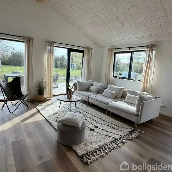 En lys stue med en hvid sofa og små borde. Sofaen står på et lyst tæppe, omgivet af store vinduer med udsigt til en terrasse og have.