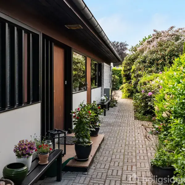 En stiforlagt terrasse af fliser langs siden af et hus med store vinduer, flankeret af grønne planter og blomster i potter, omkranset af frodige buske og træer.
