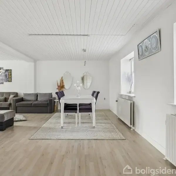 Spisestue med hvidt bord og fire stole under en moderne lampe. En stor grå sofa og en indsats ligger ved siden af. Lyse vægge, lyst trægulv og store vinduer.