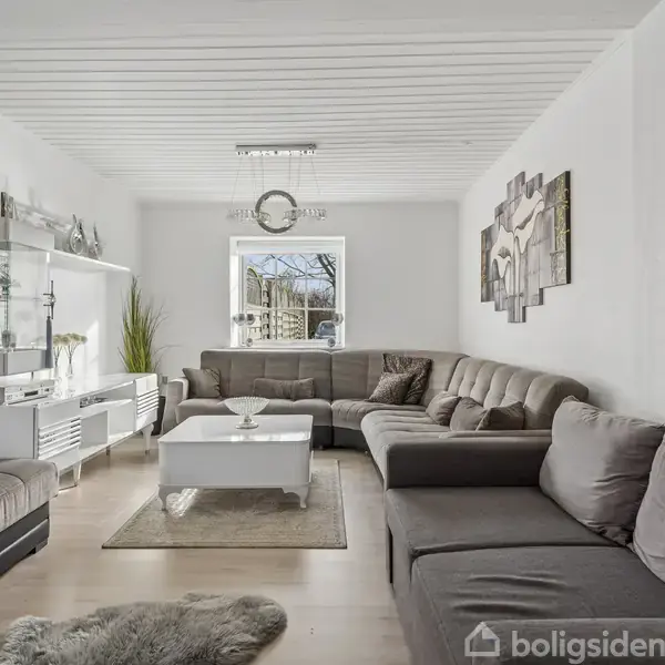 Stor, grå hjørnesofa står i en lys stue med hvidt loft, dekorativt vægkunst og sofabord. Et vindue bagved skaber en luftig atmosfære. En pænt arrangeret reol er placeret til venstre.