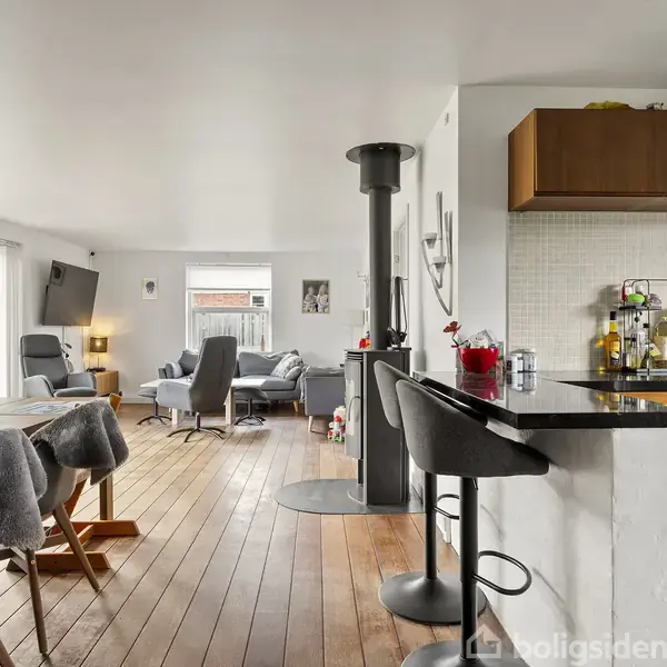 Åben stue-køkken, hvor et bord med stole og en køkkenø står på trægulv. Pejs deler rummet, sofaer og TV i baggrunden. Store vinduer giver lysindfald.