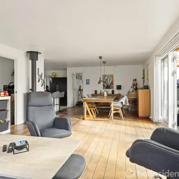 Stue med to grå lænestole og sofa omkring et bord. Børn legetøj på hylde til venstre. Åben spisestue med spisebord og stole i baggrunden. Store vinduer til højre.