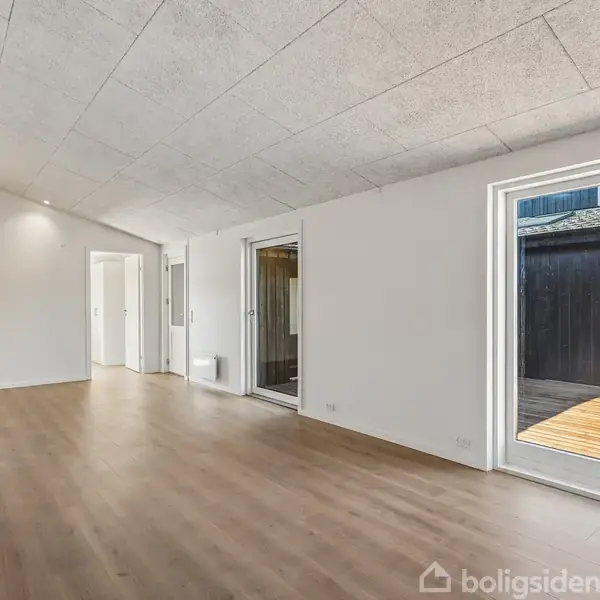 Lys stue med trægulv, hvidt loft med indbyggede spotlights. Store vinduer giver udsigt til en træterrasse udenfor. Værelse og entrédør til venstre. Ingen tekst.