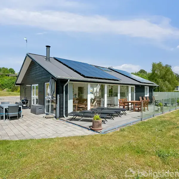 Hus med mørkt træ og solpaneler på taget står i en græsplæne. Terrasse med havebord og stole. Omgivet af grønne områder og træer under blå himmel.