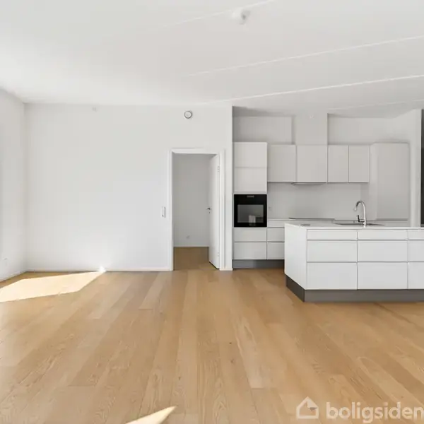 Lys stue med trægulv og et stort vindue, åben forbindelse til et moderne hvidt køkken med indbygget ovn og en ø. Rummelig og minimalistisk indretning.