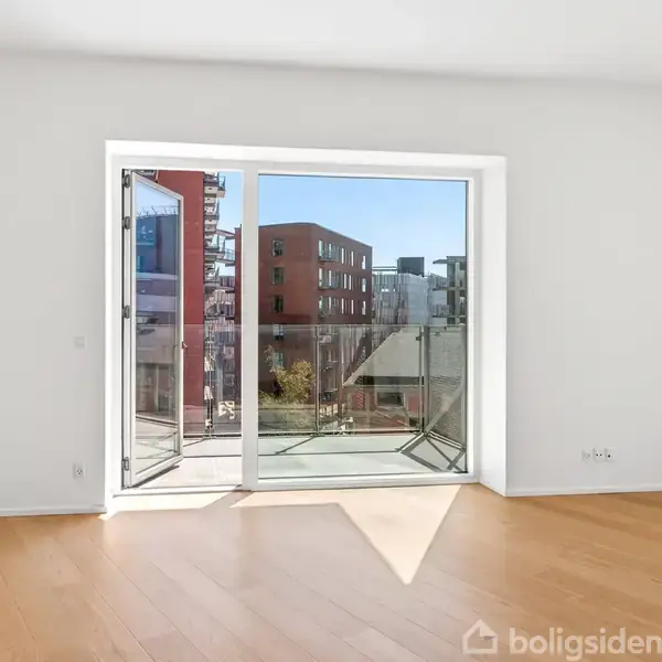 Et lyst rum med trægulv og en åben glasdør, der fører ud til en balkon. Udsigt til moderne bygninger udenfor.