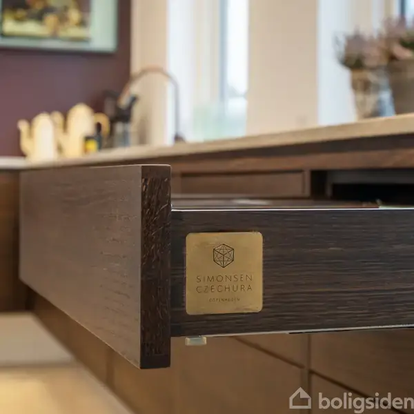 Skuffe trukket ud i et moderne køkken, med tekst "Simonsen & Czechura" på metalplade. I baggrunden ses bordplade og køkkenindretning med plantere i vindueskarmen.