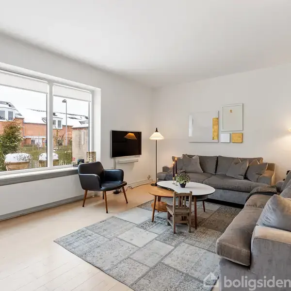 En stue med to grå sofaer omkring et rundt sofabord. En lampe er tændt bagved sofaerne. Store vinduer viser en udsigt til sne udenfor.