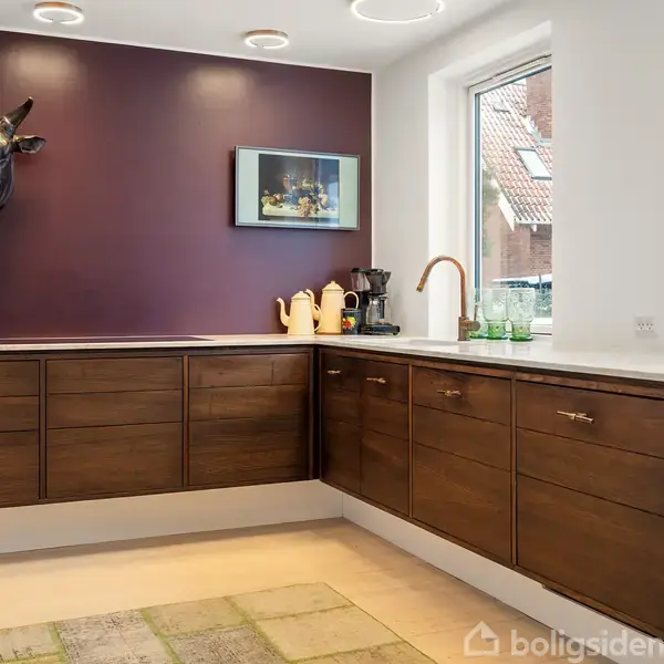 Køkken med mørkebrune skabe langs to vægge, dekoreret med en kohoved-skulptur og et billede på en lilla væg. Lysindfald fra stort vindue, flisegulv med tæppe.