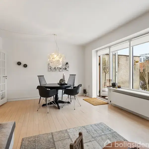 Spisebord med fire stole står i et lyst rum med trægulv, store vinduer og en sofa i forgrunden. En dør med glaspartier fører til et andet rum.