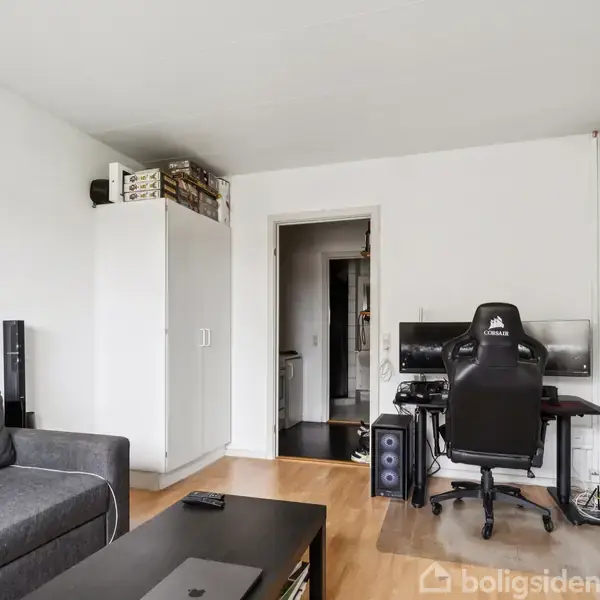 Side view af en stue med en grå sofa til venstre. Et skrivebord med en computerskærm og gamingstol står i midten. Et hvidt skab står ved siden af en dør.