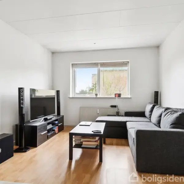 Gråt L-formet sofa i stue, står på trægulv ved tv-bord og fladskærm. Et skrivebord med stol er også til stede. Et vindue giver naturligt lys bag sofaen.