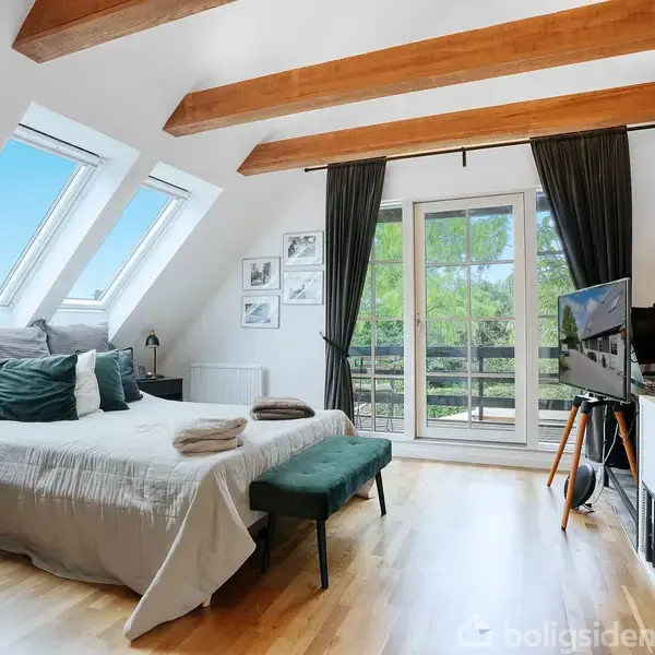 Soveværelse med seng, aktiveret af dagslys fra store ovenlysvinduer. Rummet har træbjælker, skrivebord, hylder, fladskærms-TV og dør til balkon med udsigt til træer og blå himmel.