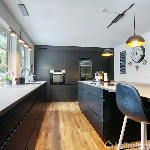 Et moderne køkken med sorte skabe langs væggen, indeholdende en ovn og køleskab. Trægulv dækker området. En stor køkkenø med barstol er i centrum, omgivet af pendellamper.