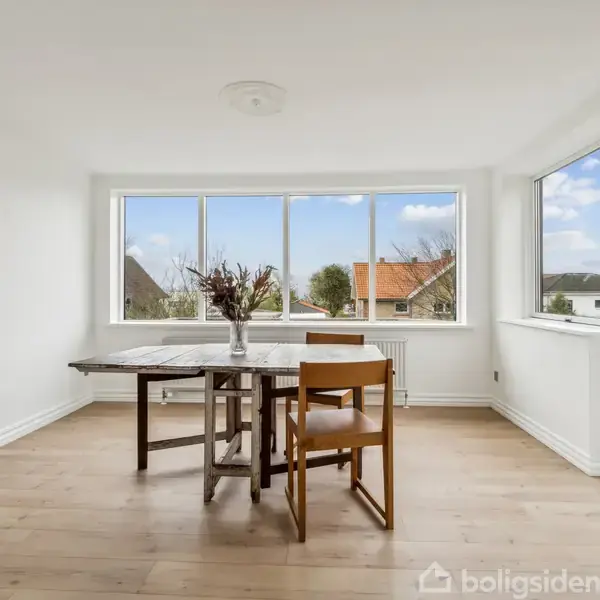 Et træbord står stille, flankeret af to stole, i et lyst rum med store vinduer. En vase med blomster pryder bordet. Udsigt til et boligområde udenfor.