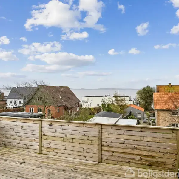 Træterrasse med udsigt, omgivet af lave bygninger og huse med farverige tage, beliggende tæt på vandet under en blå himmel med spredte skyer.