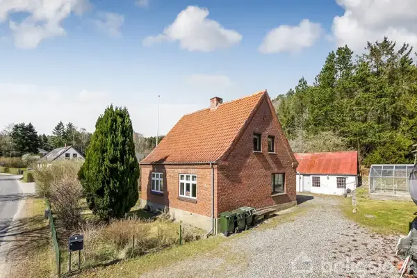 Et rødt murstenshus med orange tegltag står ved en vej. Foran huset er der en grusindkørsel og en stor grøn busk. Omkring huset er der skov og andre huse.