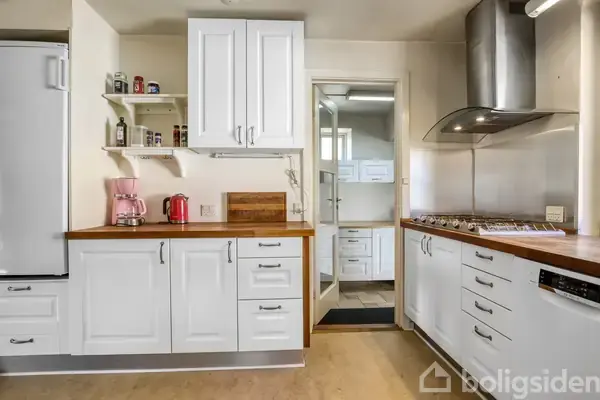 Hvidt køkken med træborde, kaffemaskine og elkedel på bordet. Komfur med fem gasblus og emhætte. Hylder med krydderier, skærebræt. Dør leder til andet rum.