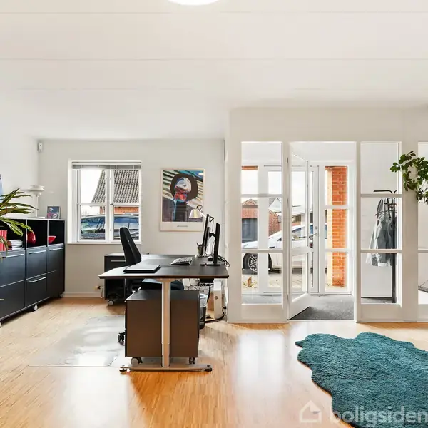 Kontor med skrivebord og stol står nær et vindue, omgivet af planter i krukker. Et åbent glasparti fører til en lys gang. Gulvtæppe giver farve på trægulvet.