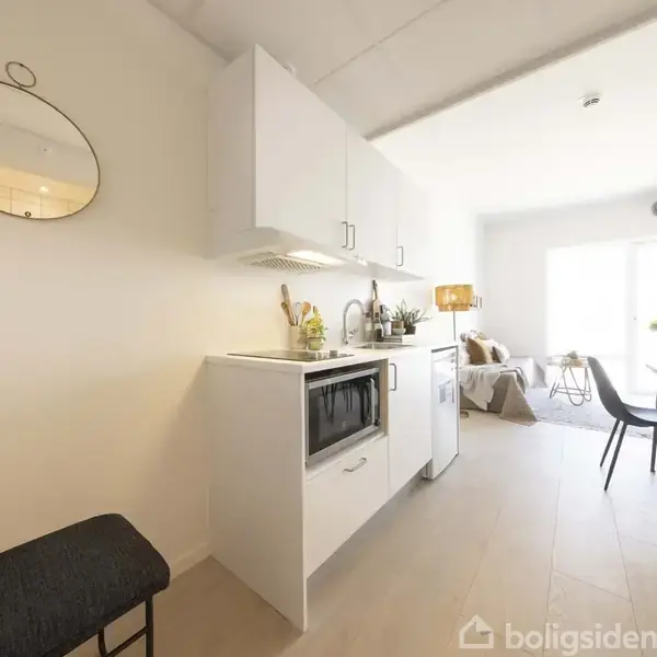 Et moderne køkken med hvide skabe og en mikroovn står i forgrunden. Baggrunden viser en lys stue med en sofa og spisebord ved store vinduer. En rund vægspejl hænger til venstre.