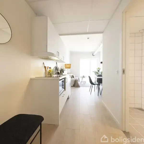 Et lyst køkken med hvide skabe og moderne apparater langs en væg. Til højre ses et badeværelse med en hængende plante. I baggrunden er en spiseplads.