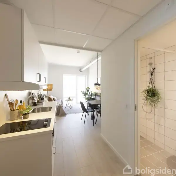 Et moderne køkken med hvidt interiør står stille, omgivet af en spisestue i baggrunden. Til højre ses en flisebeklædt badeværelse med en vask og hængende planter.