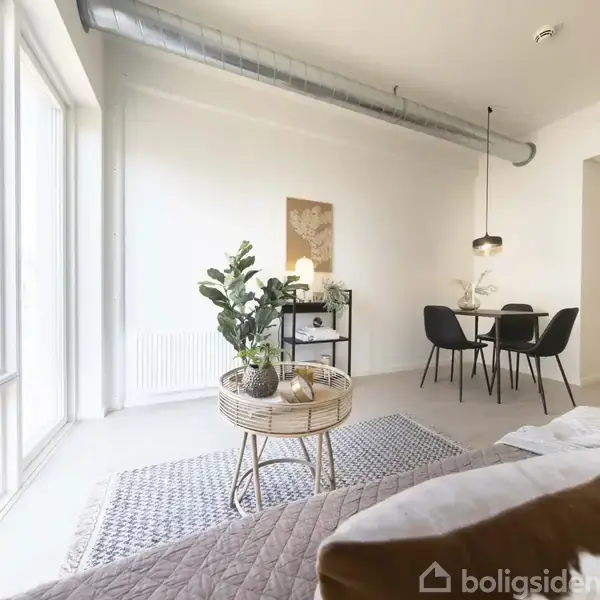 En lys stue har en sofa med puder i forgrunden. Et rundt bord bærer grønne planter. Spisebord med sorte stole ses i baggrunden. Store vinduer skaber rummelighed.
