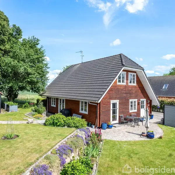 Et rødmalet hus med mørkt tag står i en velplejet have med blomsterbede. Et udendørs bord og stole er placeret på terrassen. Baggrunden har træer og en blå himmel.