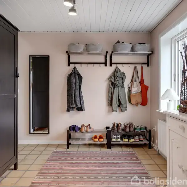 En entre har en sort garderobe, spejl, og knagerække med frakker og tasker. Skohylde med sko står nedenunder. Et vindue og en hvid kommode med dekorationer er til højre.