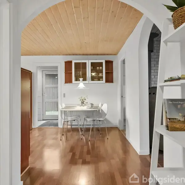 En hvid stol står ved et rundt spisebord med en hvid lampe hængende over. I baggrunden er der et træloft, et skab og døråbninger.