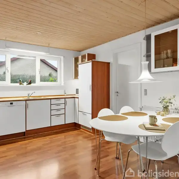 Et lyst køkken med træloft og trægulv har hvide skabe og apparater. Et rundt spisebord med fire hvide stole står ved en væg. Lys kommer ind fra et stort vindue.