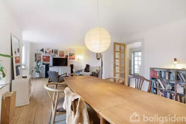 Et langt spisebord er centralt placeret med stole omkring. Over bordet hænger en rund papirlampe. I baggrunden ses en stue med sofa, planter, bogreol og et fjernsyn. Plakater hænger på væggen.