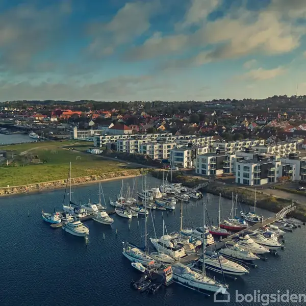 Sejlbåde ligger forankret i en marina langs en kystby. Moderne boligblokke og små huse strækker sig langs kysten under en delvist skyet himmel.
