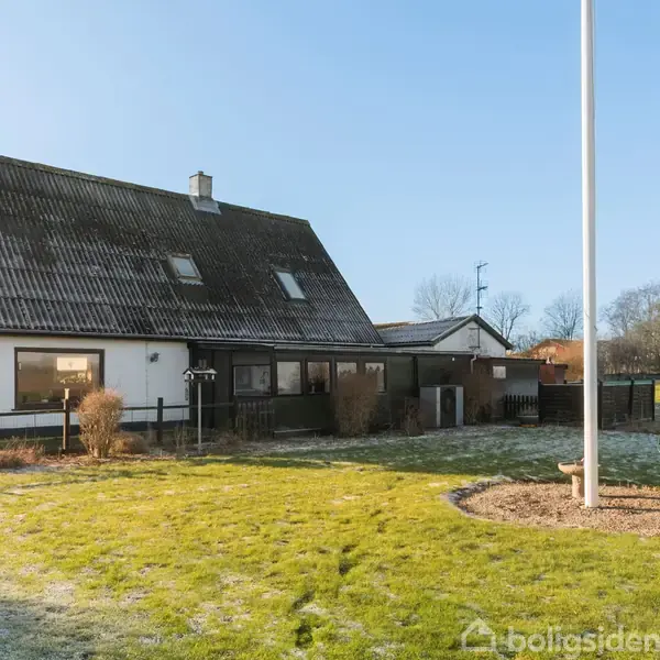 Et hus med mørkt tag, hvide vægge og store vinduer står i en velholdt have. Flagstang og græsplæne findes foran huset, omgivet af åbent landskab og nogle få træer.