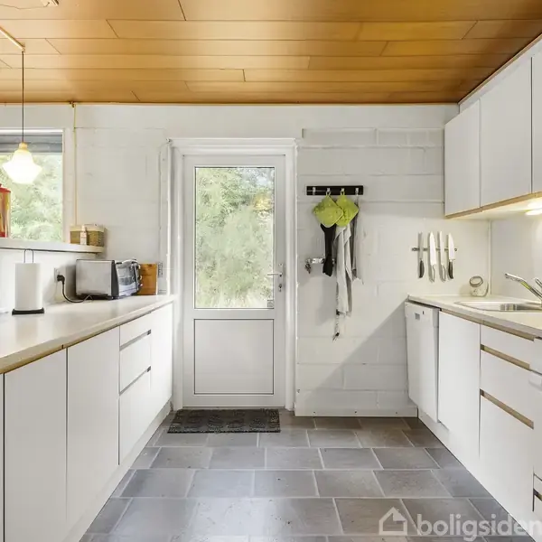 Et lyst køkken med hvide skabe og træloft. Køkkenmaskiner står på bordpladen, og knive hænger på væggen. En dør fører til en grøn have udenfor.