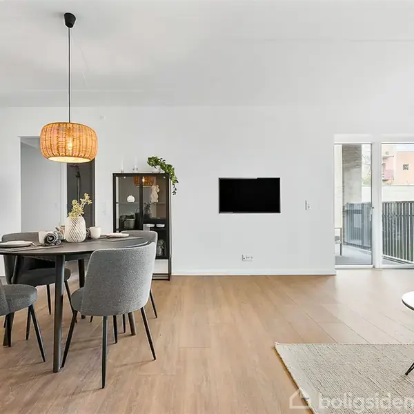 Et moderne opholdsrum med runde grå stole rundt om et spisebord. Elegant lampe hænger over bordet. Køkken i baggrunden. Store vinduer og dør til balkon til højre.