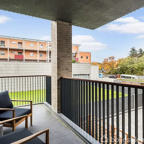 En balkon med to mørkeblå havestole står stille. Den omkranses af et rækværk, og i baggrunden ses en parkeringsplads og bygninger, omgivet af efterårsfarvede træer.