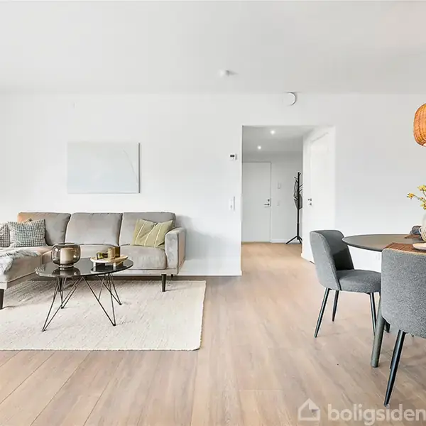 En stue med en grå sofa og sofabord placeret på et tæppe, en gulvlampe ved siden af. Rundt spisebord med grå stole og en hængende lampeskærm over.