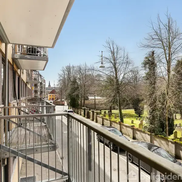En balkon med metalrækværk strækker sig ud fra en murstensbygning. Fortovet og biler ses nedenfor, mens en kirkegård med gravsten og træer ligger på den anden side af vejen.